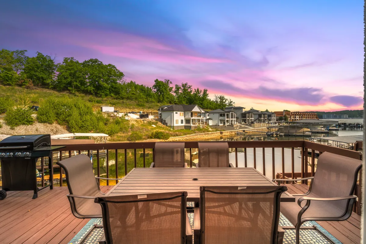 The image showcases an outdoor dining area with a table and grill set against a serene waterfront backdrop. Gentle lake waters and distant houses create a peaceful atmosphere, inviting guests to relax, share meals, and enjoy the sunset. The setting is perfect for social gatherings and unwinding by the lake.