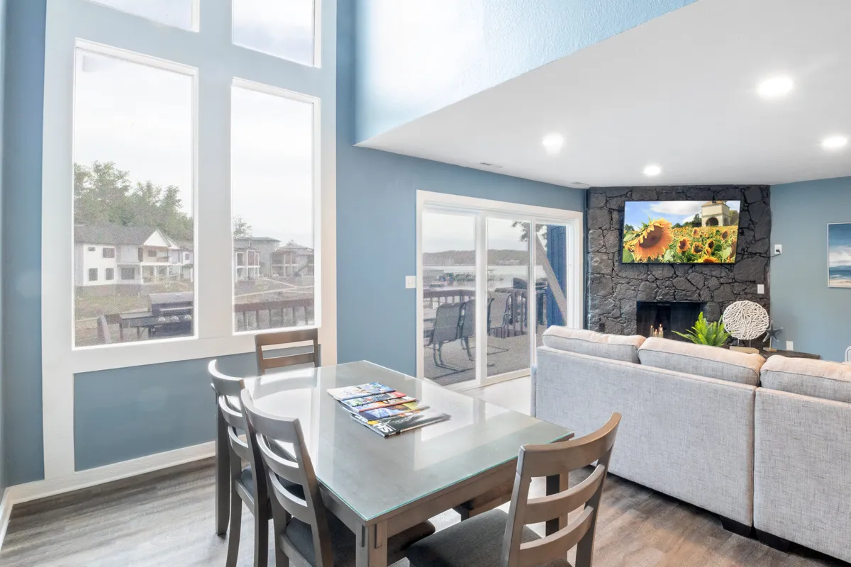 A bright, inviting living and dining area featuring a comfortable sofa, a dining table with multiple chairs, a welcoming fireplace, and a large TV. Expansive windows provide a scenic backdrop, filling the space with natural light and offering beautiful lake views.