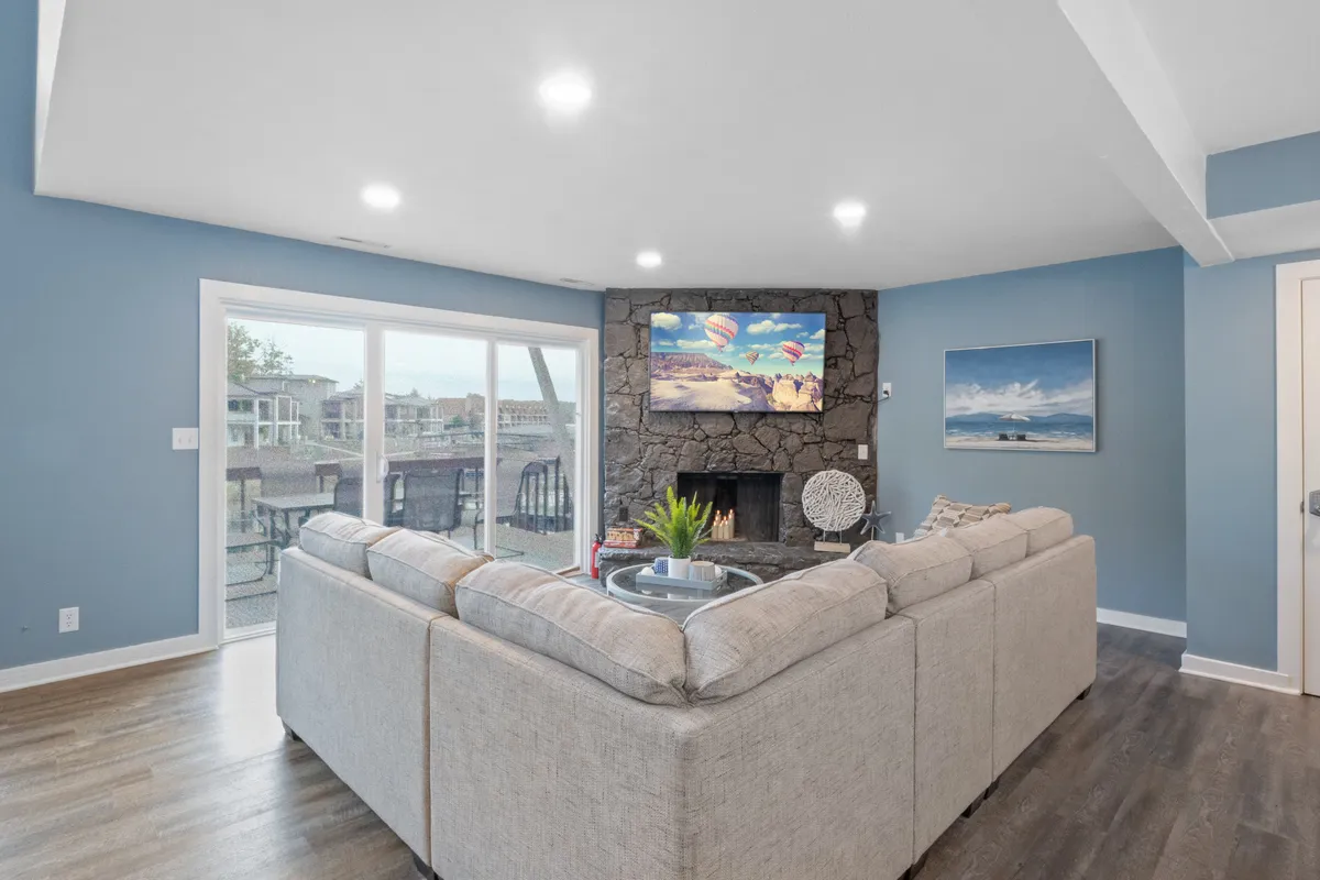 A spacious open-concept living room featuring a large sectional sofa positioned around a stone fireplace. Large windows allow natural light to filter in, offering serene views of the cove. A flat-screen television is mounted above the fireplace, creating a cozy entertainment hub ideal for relaxing or gathering with family and friends.