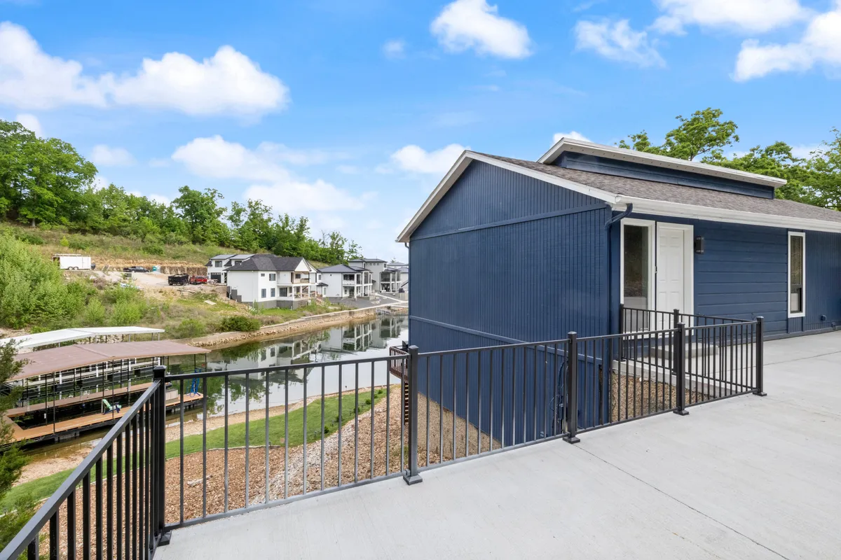 This image captures the blue exterior of the house, a patio area with a railing overlooking the water, lush greenery, and docked boats nearby. The calm water and inviting outdoor space suggest a tranquil lakeside setting ideal for families.