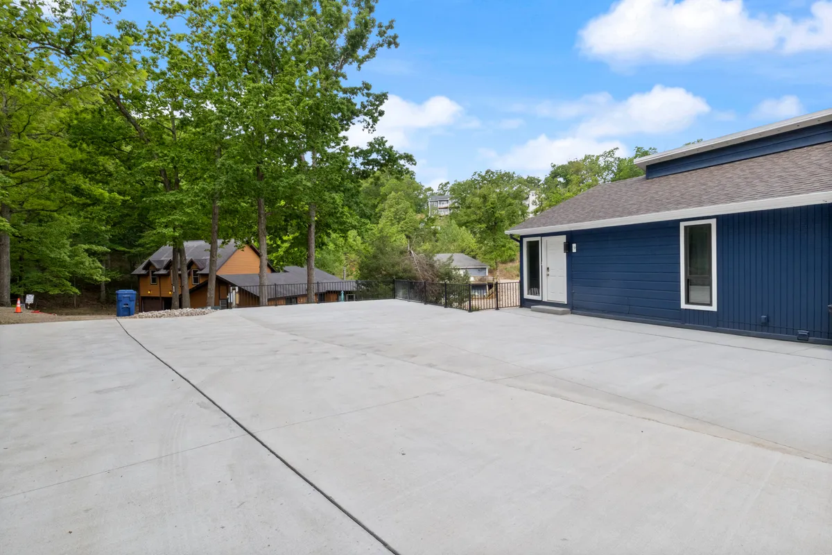 This exterior view shows the blue house amid trees, a concrete driveway, and neighboring homes set at a comfortable distance. The serene surroundings and natural setting emphasize the property's placement in a quiet cove, suggesting access to nature and outdoor relaxation—perfect for couples looking to unwind or take romantic walks.