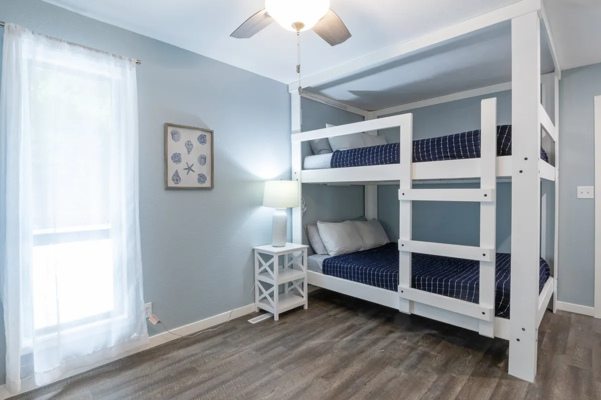 A playful yet comfortable bedroom featuring a bunk bed, nightstand with lamp, framed artwork, and a window dressed with sheer curtains. The room exudes a welcoming vibe, ideal for children or families.