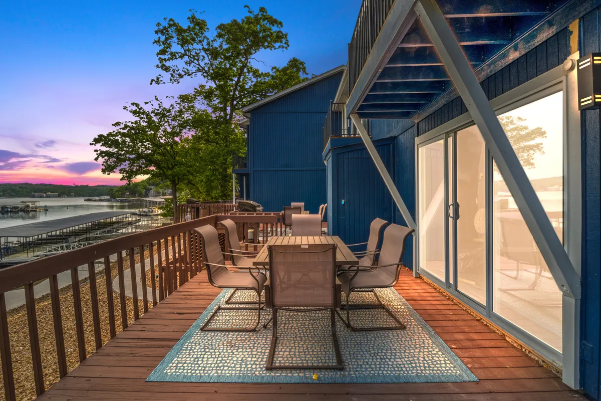 An outdoor dining table set on a deck with surrounding trees and a stunning sunset over the water. The scene suggests friendly gatherings, al fresco meals, and relaxation close to nature.