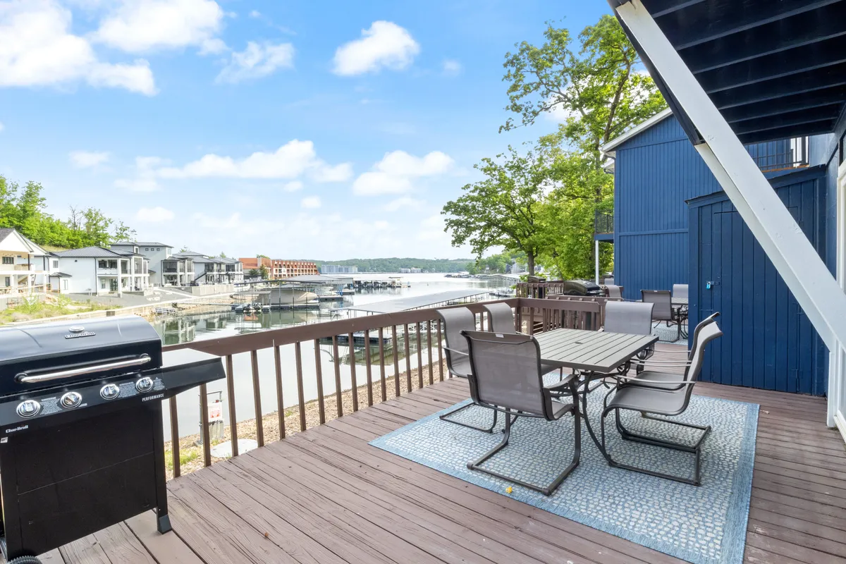 A waterfront patio with an outdoor dining table, grill, trees, and boats visible in the marina. The scene invites groups to gather for meals, barbecue, and celebrate while taking in the lakeside atmosphere.