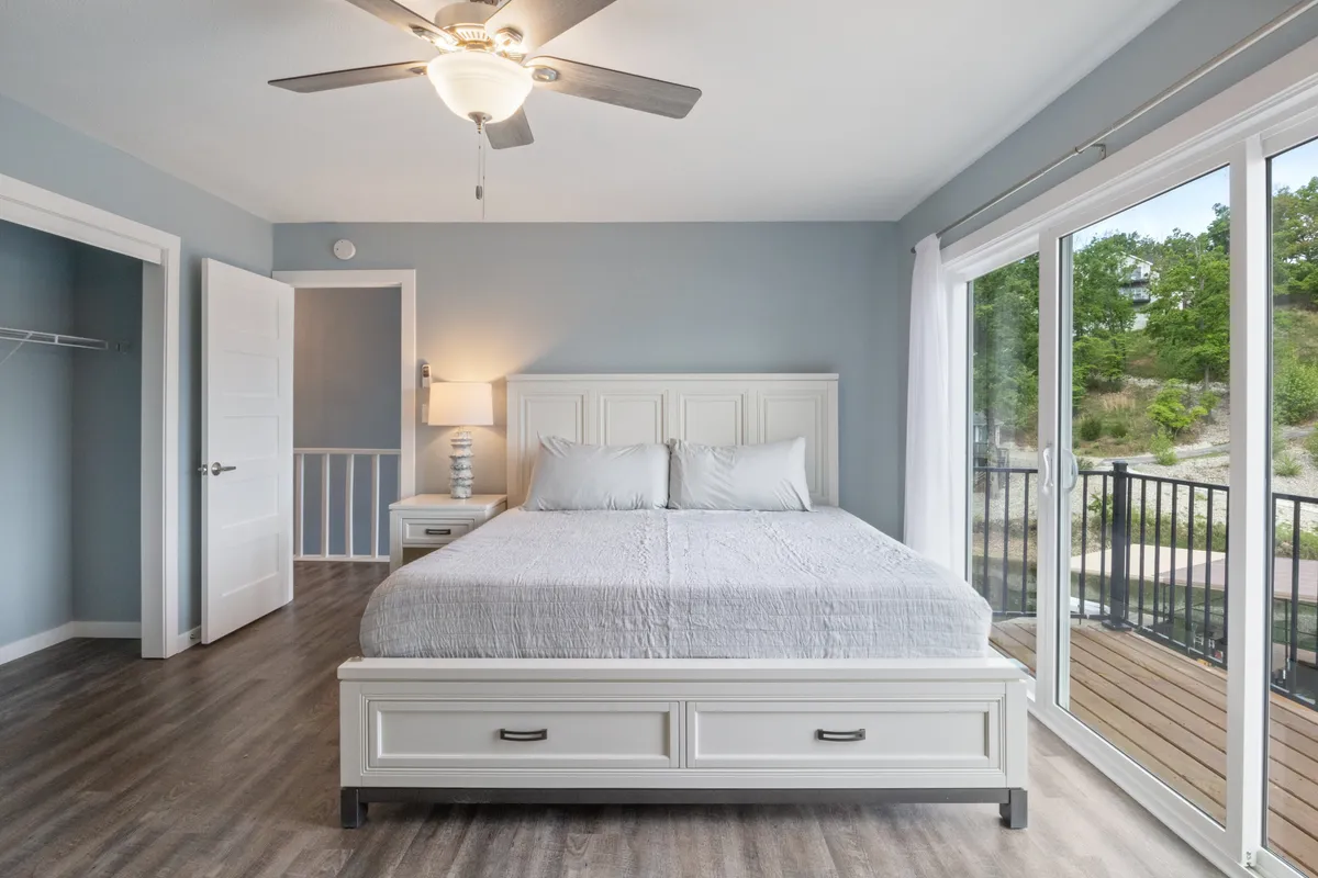 A tranquil bedroom with a neatly made bed positioned next to large windows, a bedside lamp casting a warm glow, and soothing neutral-toned decor. Sunlight filters in, hinting at the promise of blackout curtains for restful sleep.