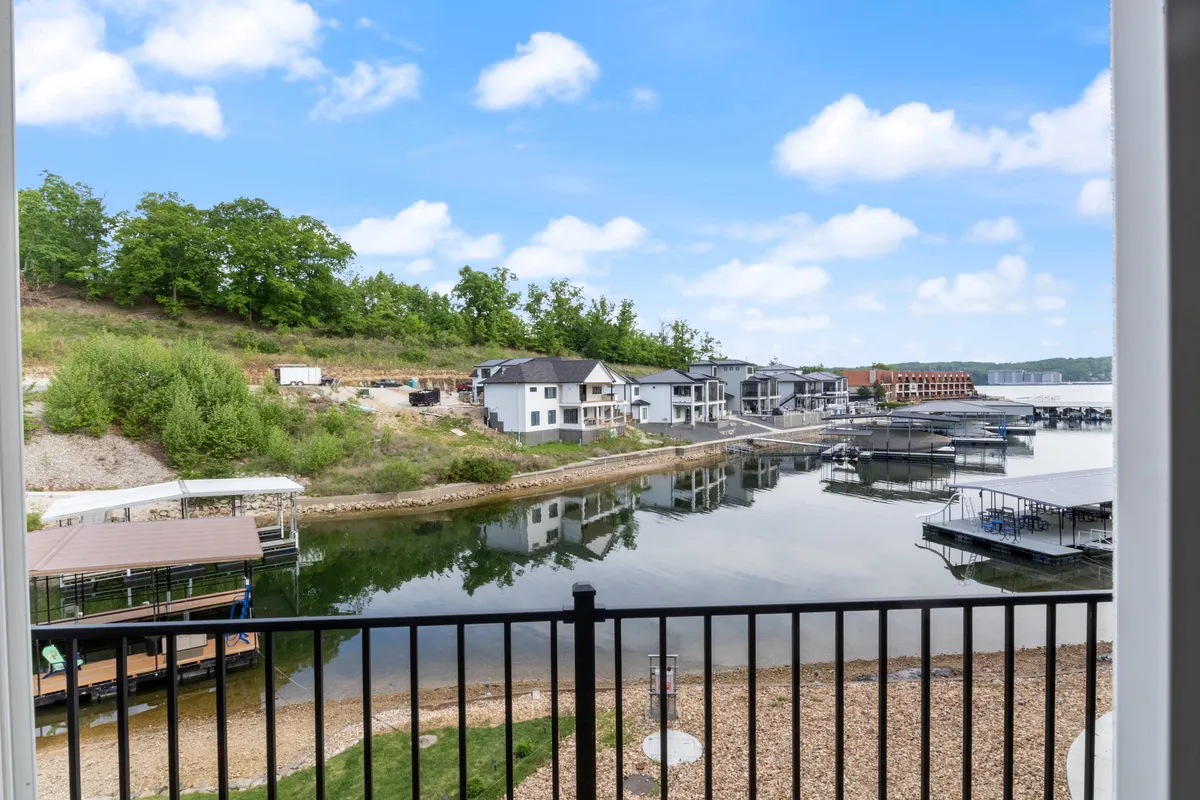A tranquil lakeside scene captures a modern dock extending over calm water, surrounded by lush greenery and distant contemporary houses. The dock features areas for seating and relaxing, with ample space for activities like fishing, boating, or simply enjoying the view.